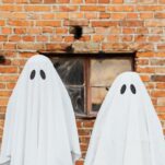 two kids dress in white sheet ghost costumes with a brick building in the background